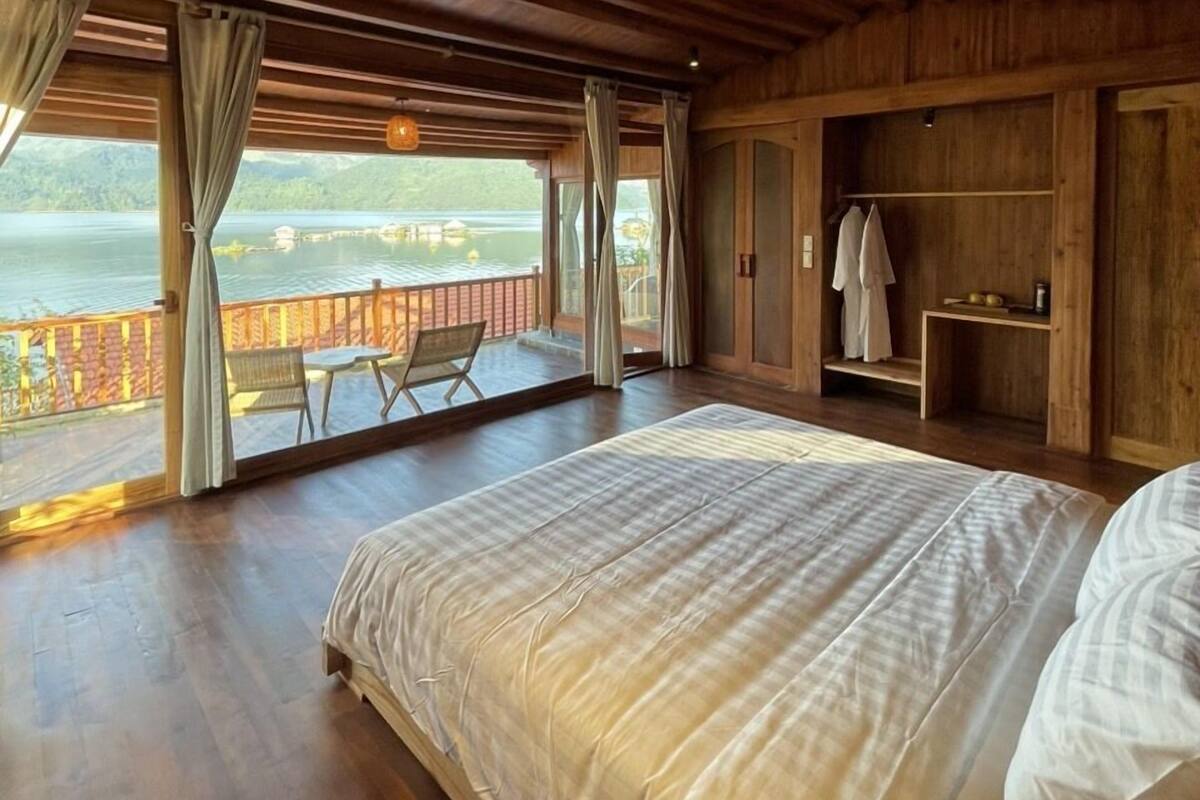 Un dormitorio amplio con una cama grande, vigas de madera en el techo, un balcón con vista al agua y una pequeña zona de estar.