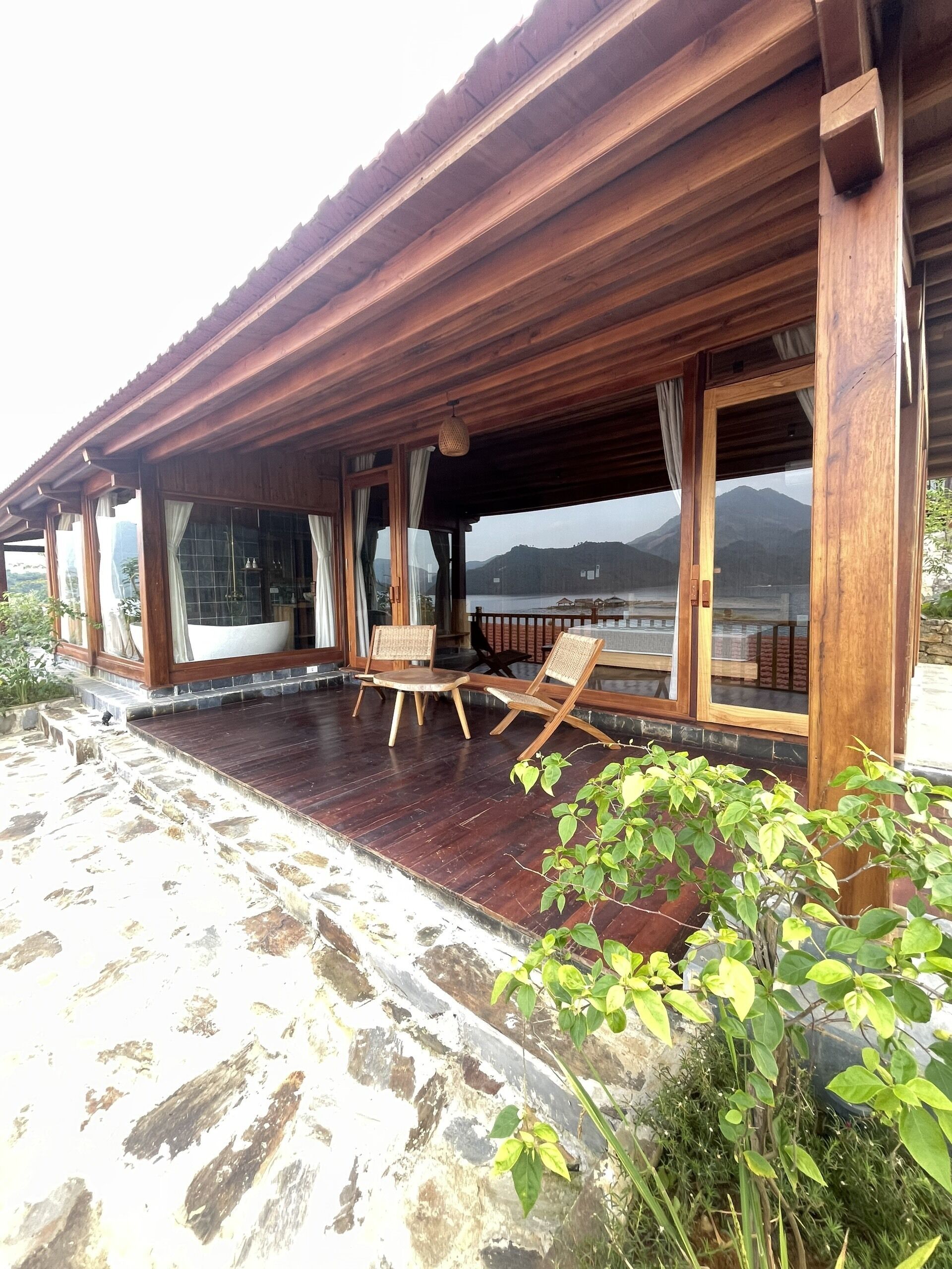 A wooden covered patio with a stone floor, large windows, and a view of a lake and mountains.