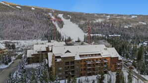Un hotel de varios pisos con un telesilla al fondo, rodeado de árboles nevados y un paisaje montañoso.