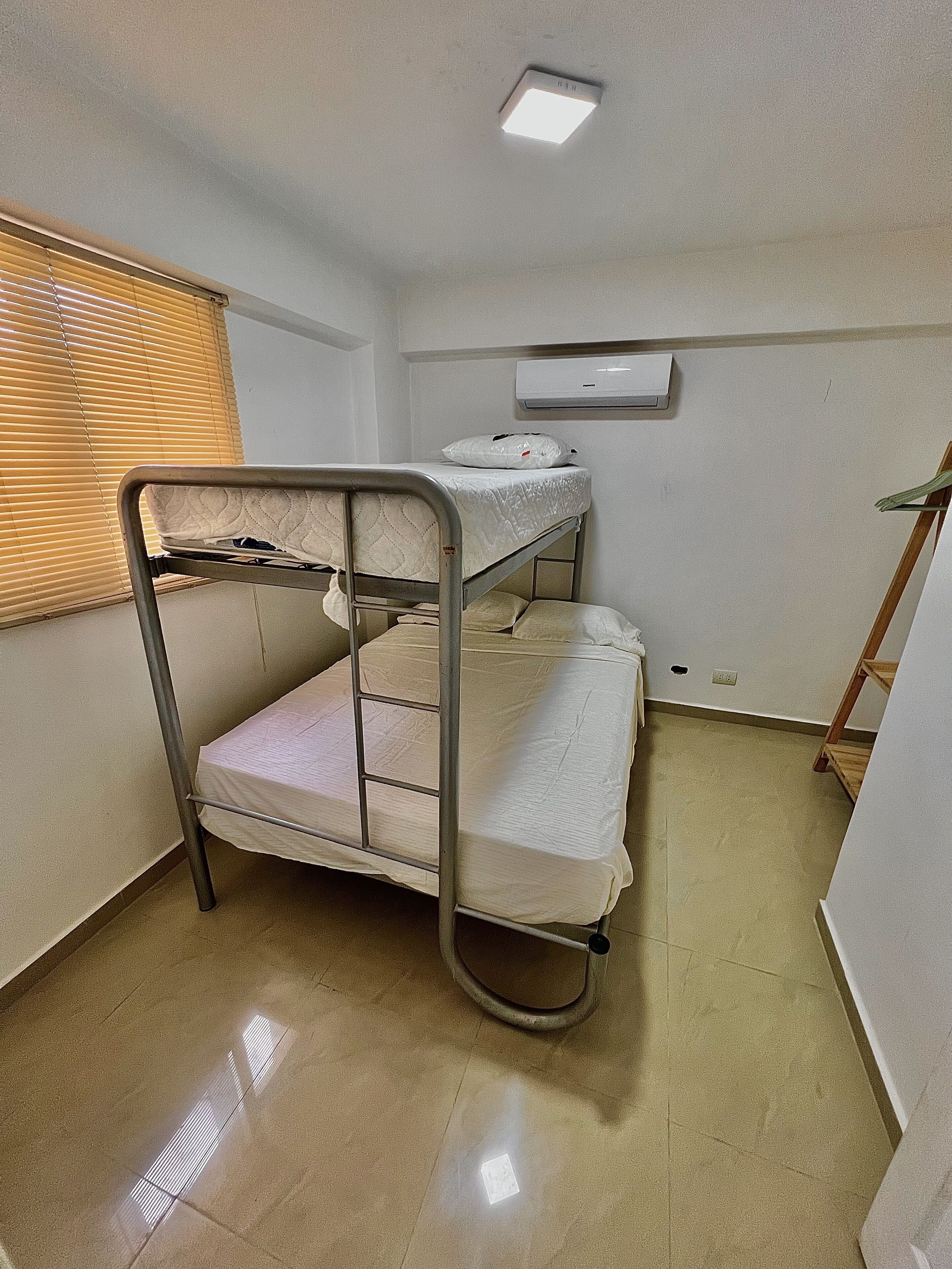 Een slaapkamer met een stapelbed, een plafondlamp, een airconditioning en een raam met zonwering.