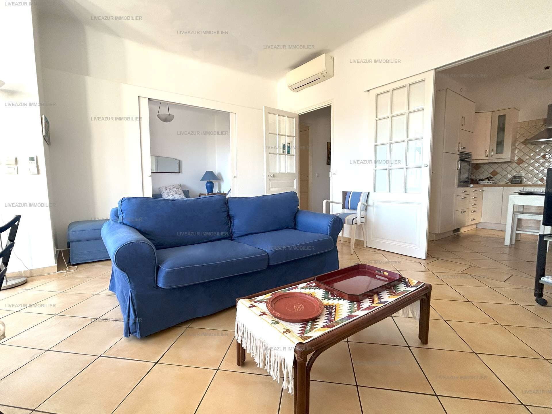 A living room with a blue sofa, a coffee table with a patterned tablecloth, and a kitchen area visible through an open door.