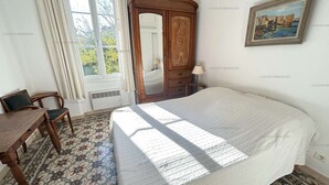 A bedroom with a bed, a wooden wardrobe, a chair, a table, and a painting on the wall.