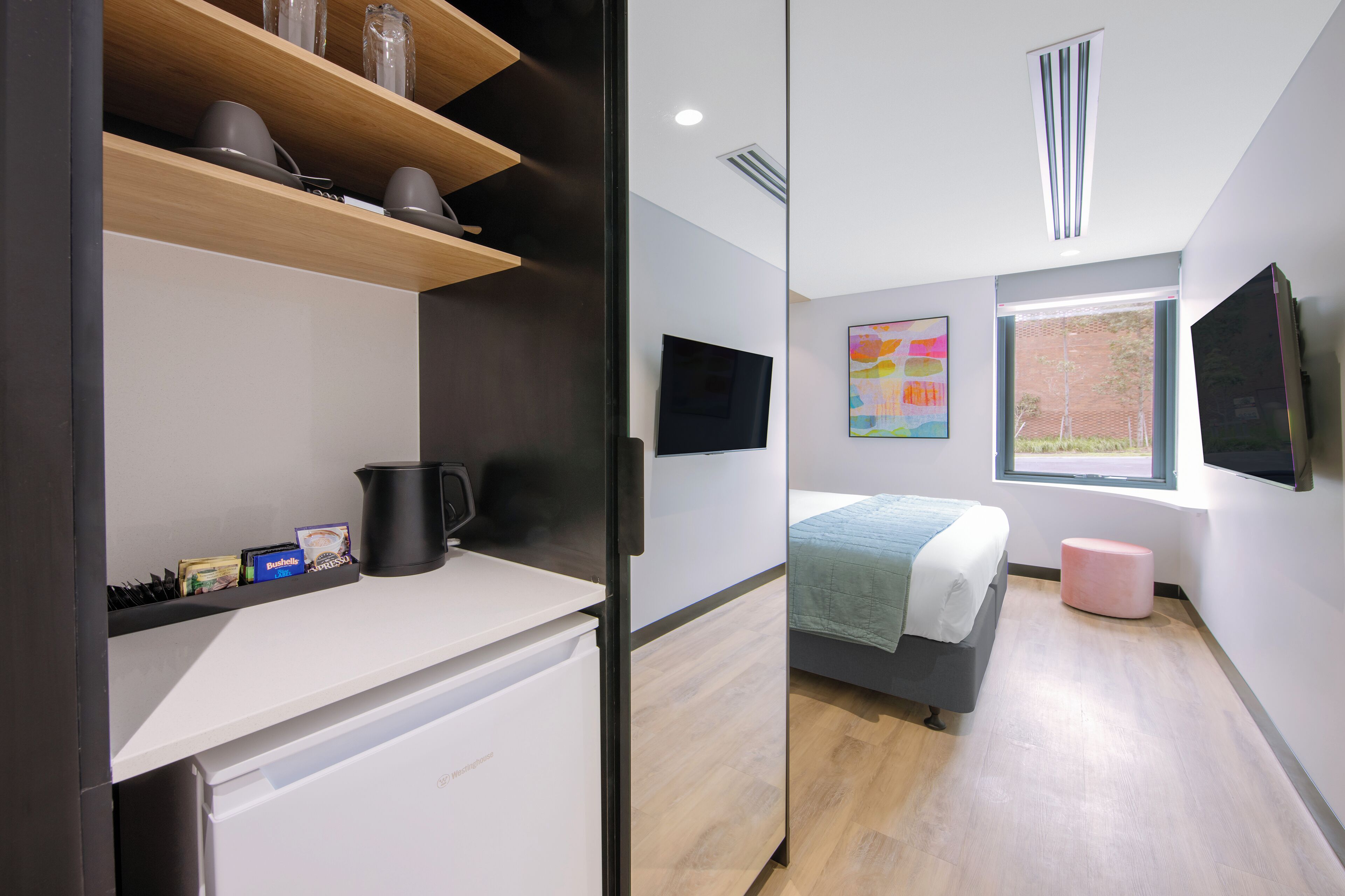 Una habitación de hotel moderna con kitchenette, una cama con manta verde, un reposapiés rosa, un ventanal grande y un cuadro abstracto colorido en la pared.