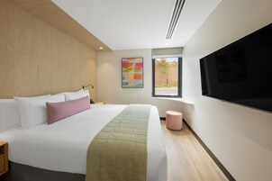 Une chambre d’hôtel moderne dotée d’un grand lit, d’un téléviseur à écran plat, d’une fenêtre offrant une vue agréable et d’un tableau abstrait aux couleurs vives accroché au mur.