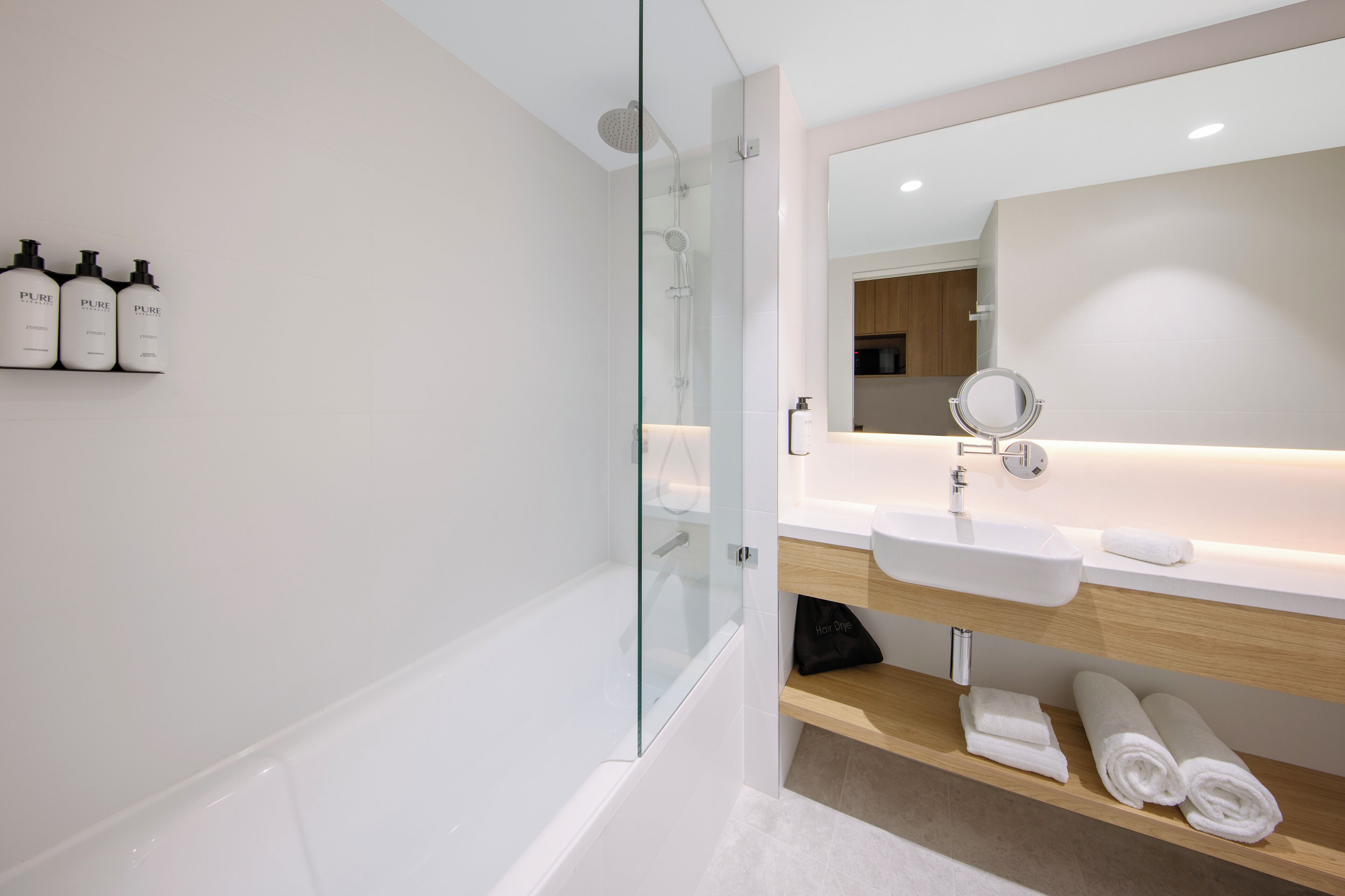 Un baño moderno con ducha de vidrio, un lavabo blanco y un espejo encima. Hay tres botellas de champú Pure en la pared.