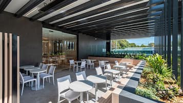 Uma área de estar ao ar livre com mesas e cadeiras brancas, um design de teto moderno e vista para vegetação e um edifício ao fundo.