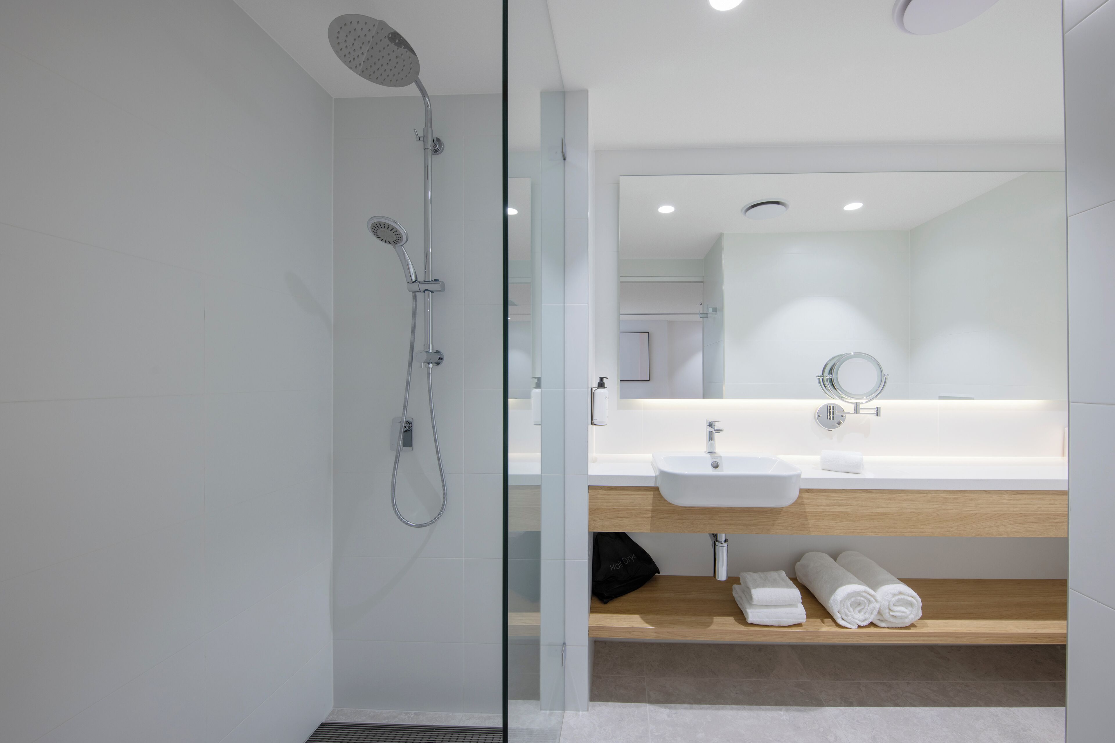 A modern bathroom with a glass shower enclosure, a white sink, and a wooden shelf with towels.