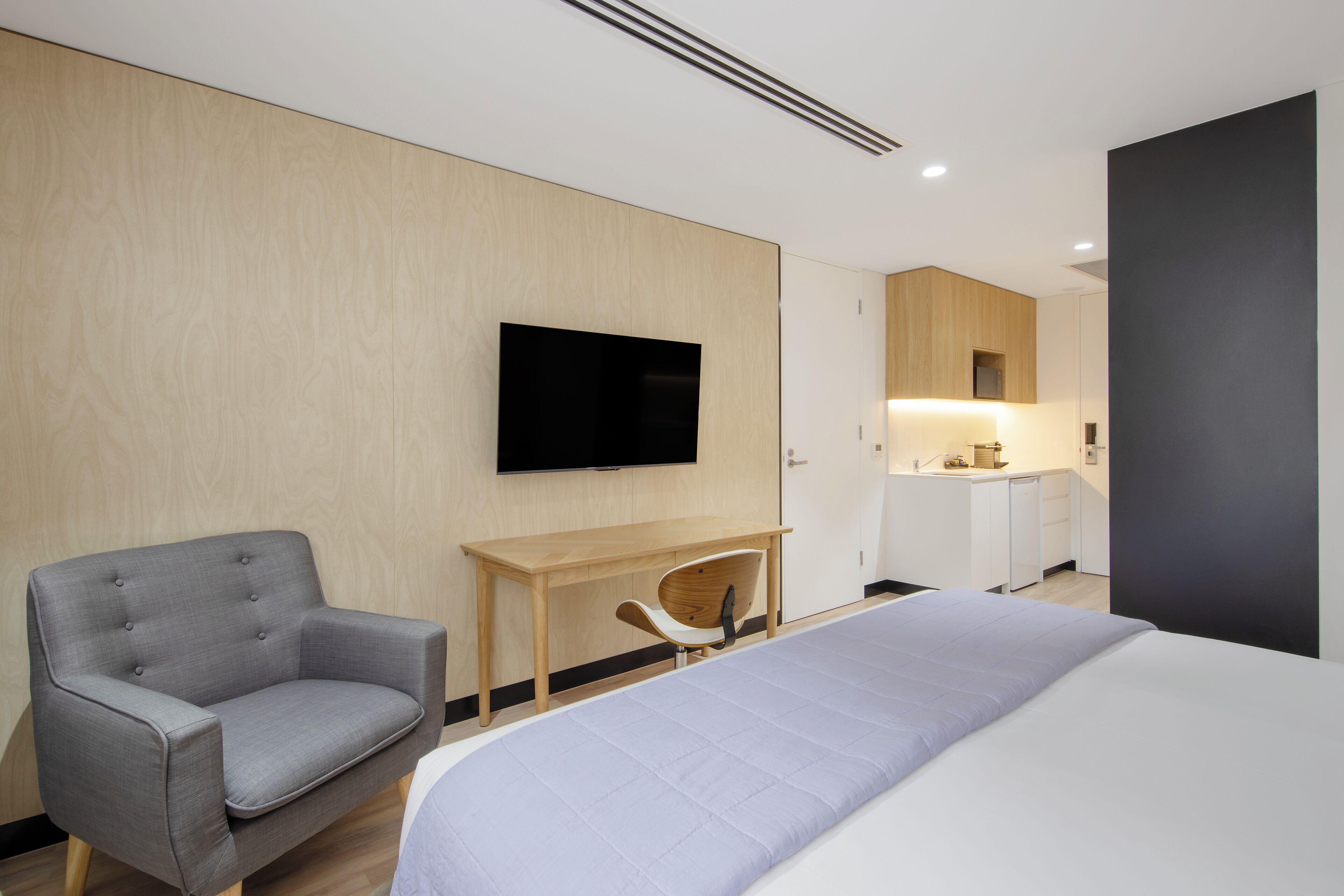 A modern hotel room with a large bed, a grey armchair, a wooden desk, a flat-screen TV, and a kitchenette in the background.
