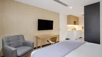 A modern hotel room with a large bed, a grey armchair, a wooden desk, a flat-screen TV, and a kitchenette in the background.