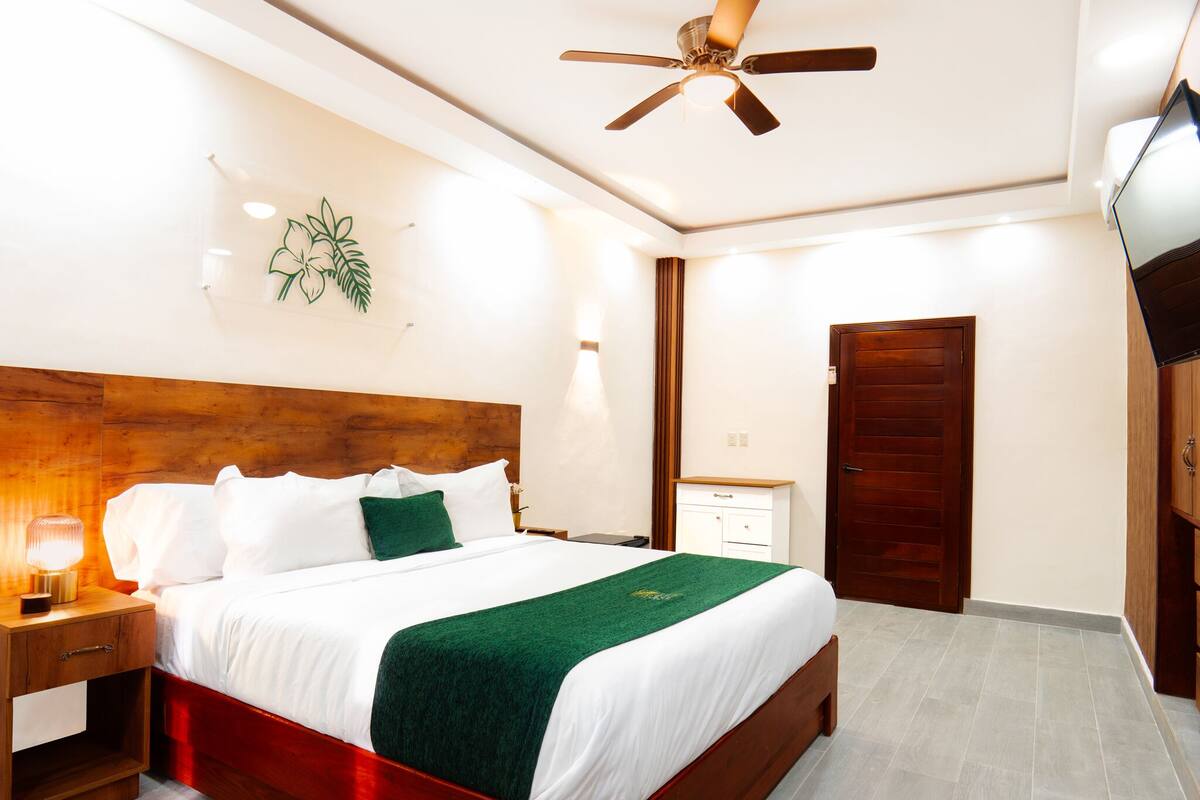 A hotel room with a wooden headboard, a bed with white linens and a green throw, a nightstand with a lamp, a ceiling fan, and a wooden door.