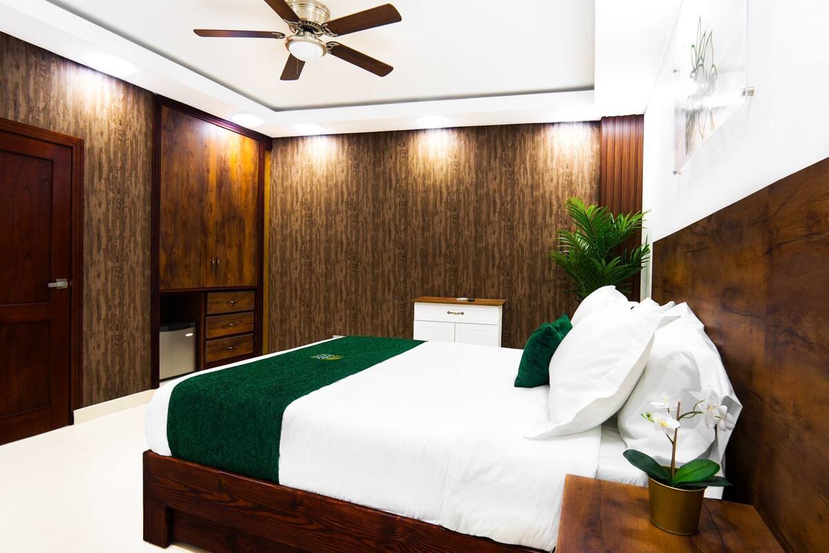 A bedroom with a wooden headboard, a bed with white and green bedding, a nightstand with a plant, and a ceiling fan.