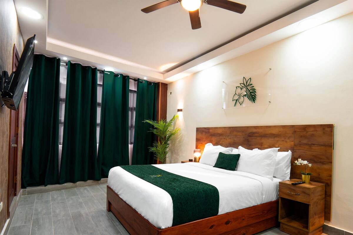 A modern hotel room with a wooden bed, green curtains, a ceiling fan, and a wall-mounted TV.