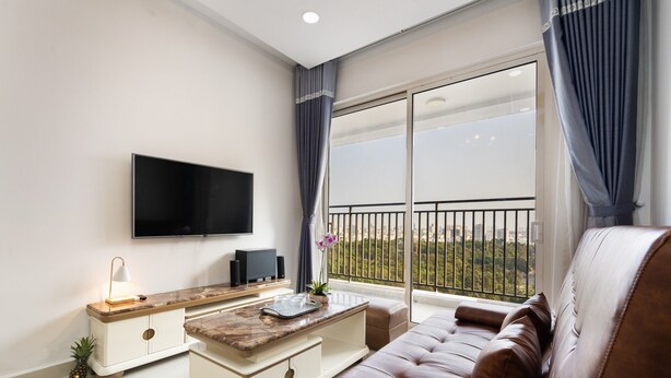 A modern living room with a flat-screen TV, a wooden coffee table, a brown leather sofa, and a balcony with a view of greenery. - Golden Mansion Apartments Phu Nhuan (Ho Chi Minh City)