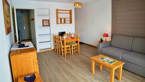A compact living space with a dining table, chairs, a sofa, a small wooden table with magazines, a lamp, and a wall-mounted shelf with a picture frame. - Studio 4 ppl, balcony, near the ski station (Les Deux Alpes)
