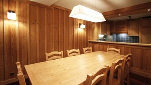 Ett rum med träpanel på väggarna, ett stort matbord och stolar, en hängande lampa och ett köksområde med skåp och en bänkskiva.