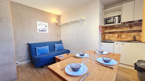 Un espacio habitable compacto con un sofá, una mesa de comedor para dos personas, una kitchenette con microondas y un refrigerador pequeño.