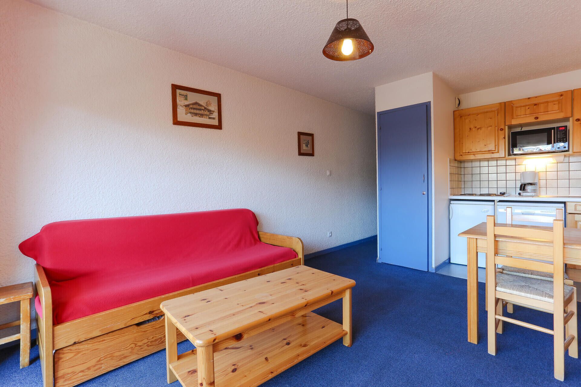 A compact living space with a red sofa, wooden table, kitchenette, and a blue door.