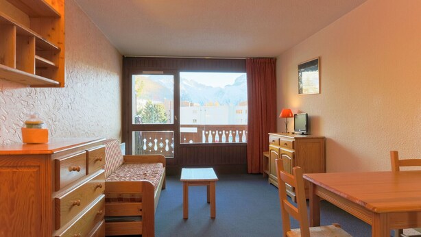 A cozy room with a wooden bed, a small table, a chair, a TV, and a window with a view of mountains. - Studio 4 ppl, splendid view from balcony (Les Deux Alpes)