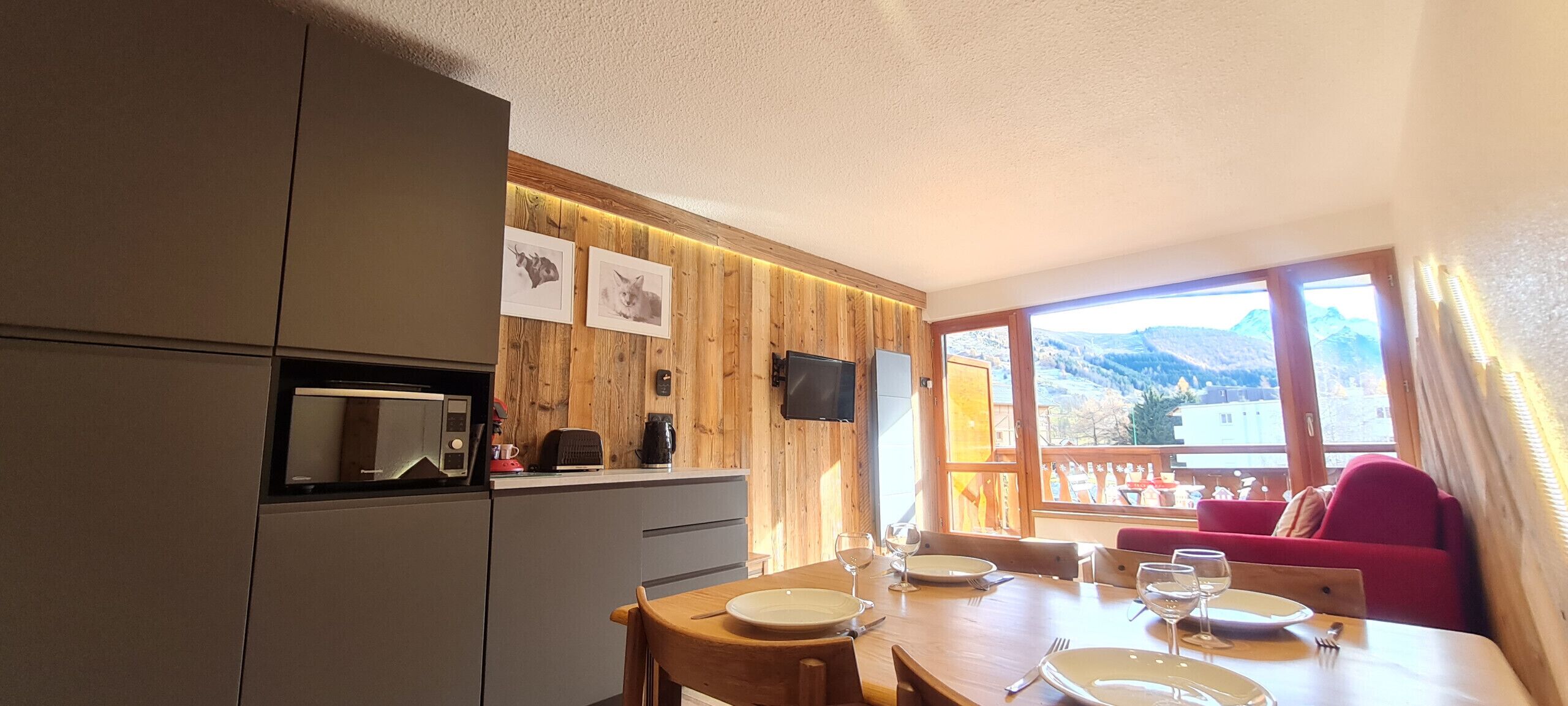 Een moderne keuken met houten lambrisering, een eethoek met een tafel gedekt voor twee personen, en een uitzicht op bergen door grote ramen.