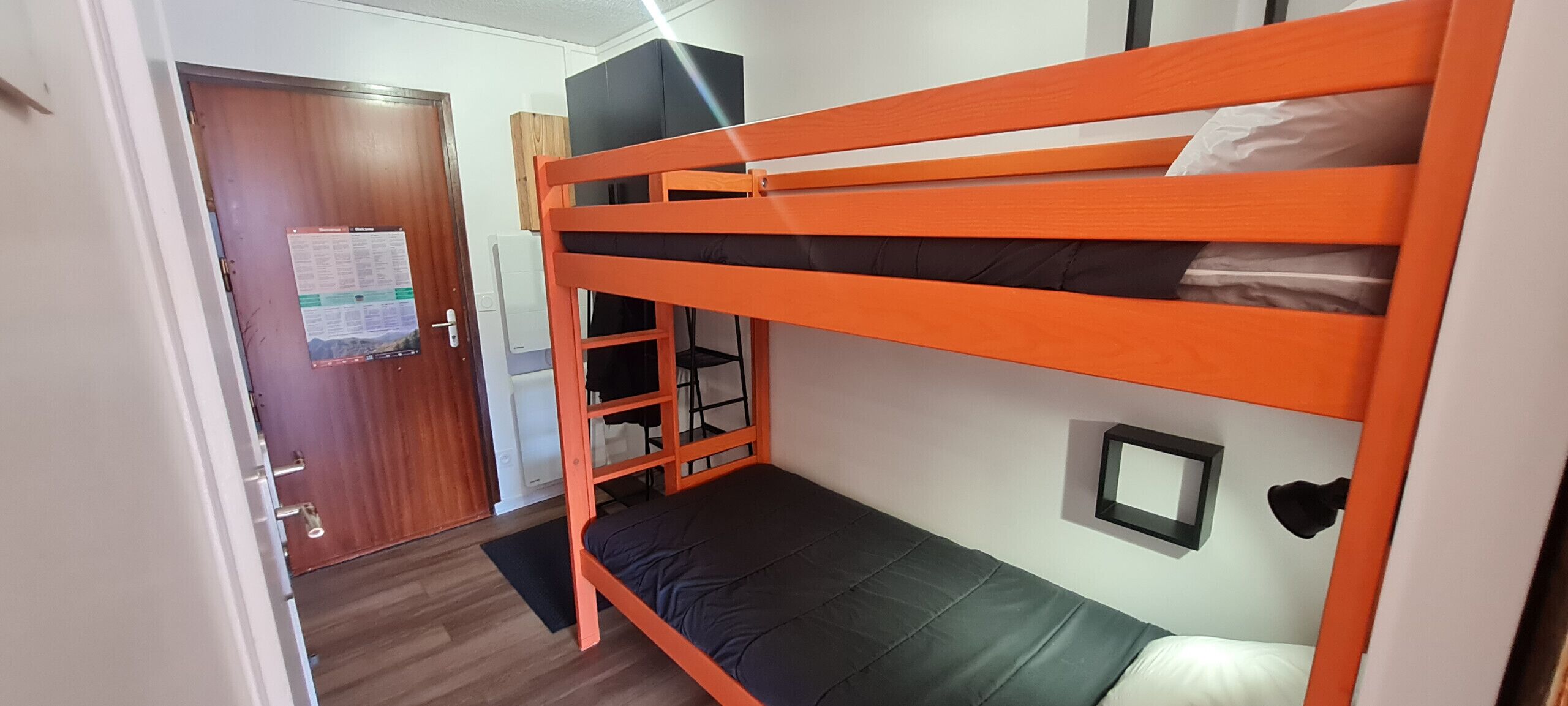 Une chambre avec un lit superposé, une porte en bois, une petite étagère et un miroir fixé au mur.