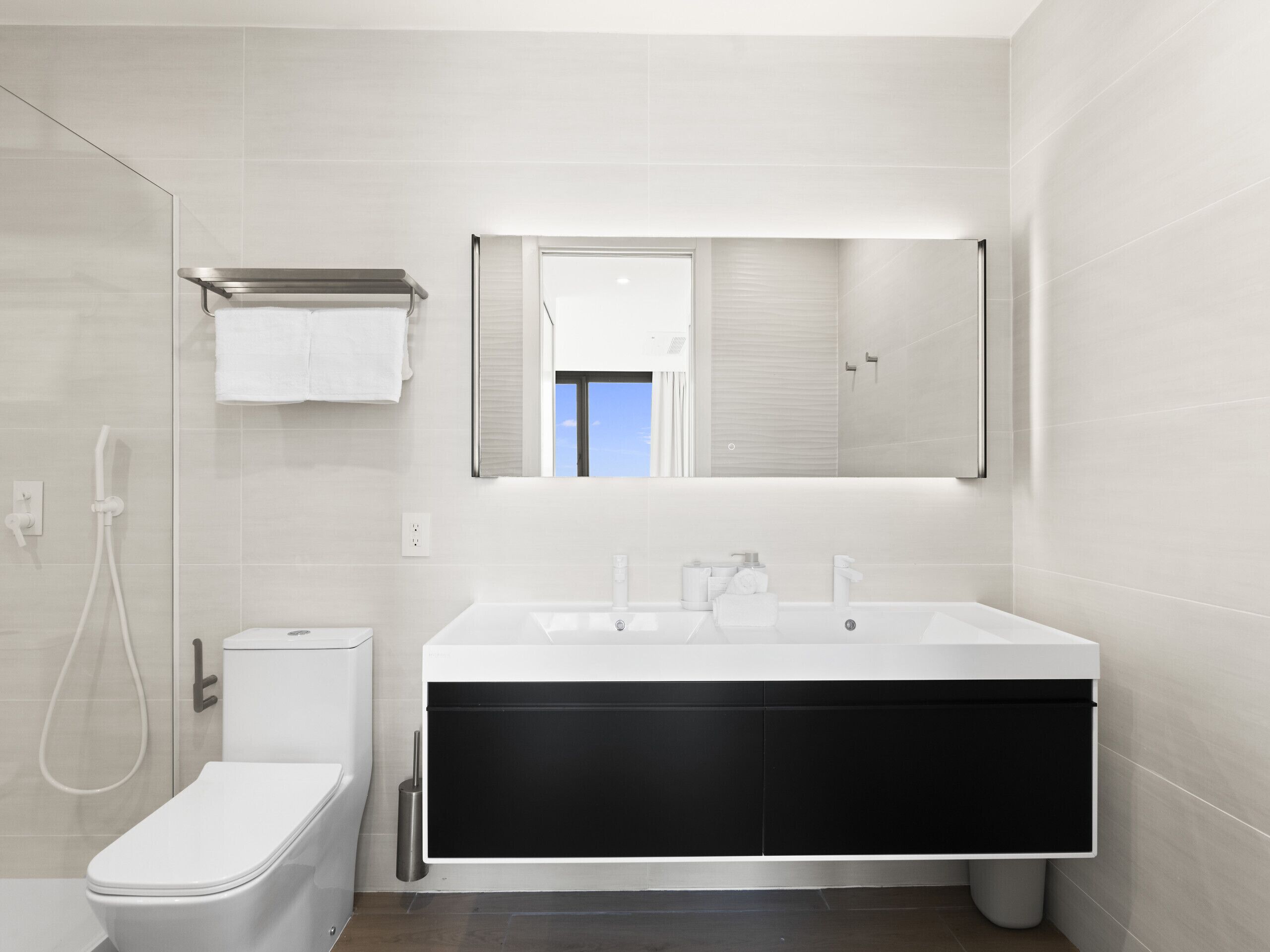 Une salle de bain moderne avec des toilettes blanches, une douche, un grand lavabo avec un miroir au-dessus et un meuble noir en dessous.
