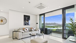 A modern living room with a sofa, coffee table, and ottoman, a large window with a view of a lake and mountains, and a ceiling with a smoke detector and ventilation. - B-1702 Stunning ocean view 2 bedroom (Cupecoy)