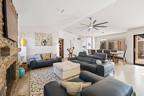 A modern living room with a fireplace, a ceiling fan, and a dining area with a large window. - Tranquil Oasis near El Paseo | Pool, Hot Tub and Sauna | Mountain Views & Outdoor Entertainment (Palm Desert)
