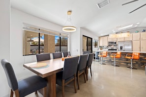 A modern kitchen with stainless steel appliances, a dining area with a wooden table and grey chairs, and a view of the outdoors through large windows. - Tranquil Oasis near El Paseo | Pool, Hot Tub and Sauna | Mountain Views & Outdoor Entertainment (Palm Desert)