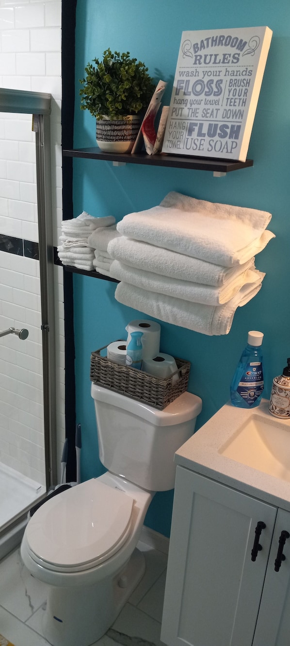 A bathroom with a toilet, a sink, and a shower. There are towels stacked on a shelf, a sign with bathroom rules, and toiletries on the sink.
