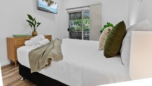 Un dormitorio ordenado con una cama, una mesita de noche con toallas, una planta en maceta y una ventana con vistas a zonas verdes.
