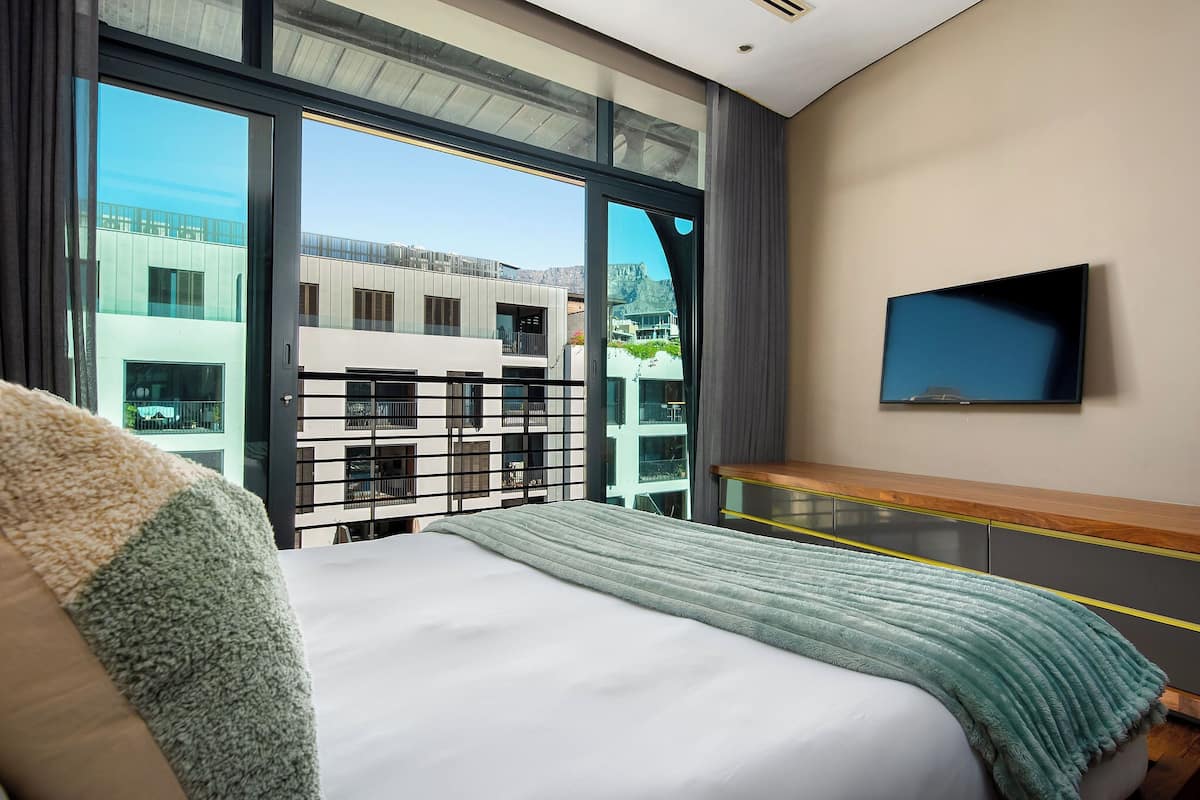 A modern hotel room with a large bed, a TV mounted on the wall, and a balcony with a view of buildings.