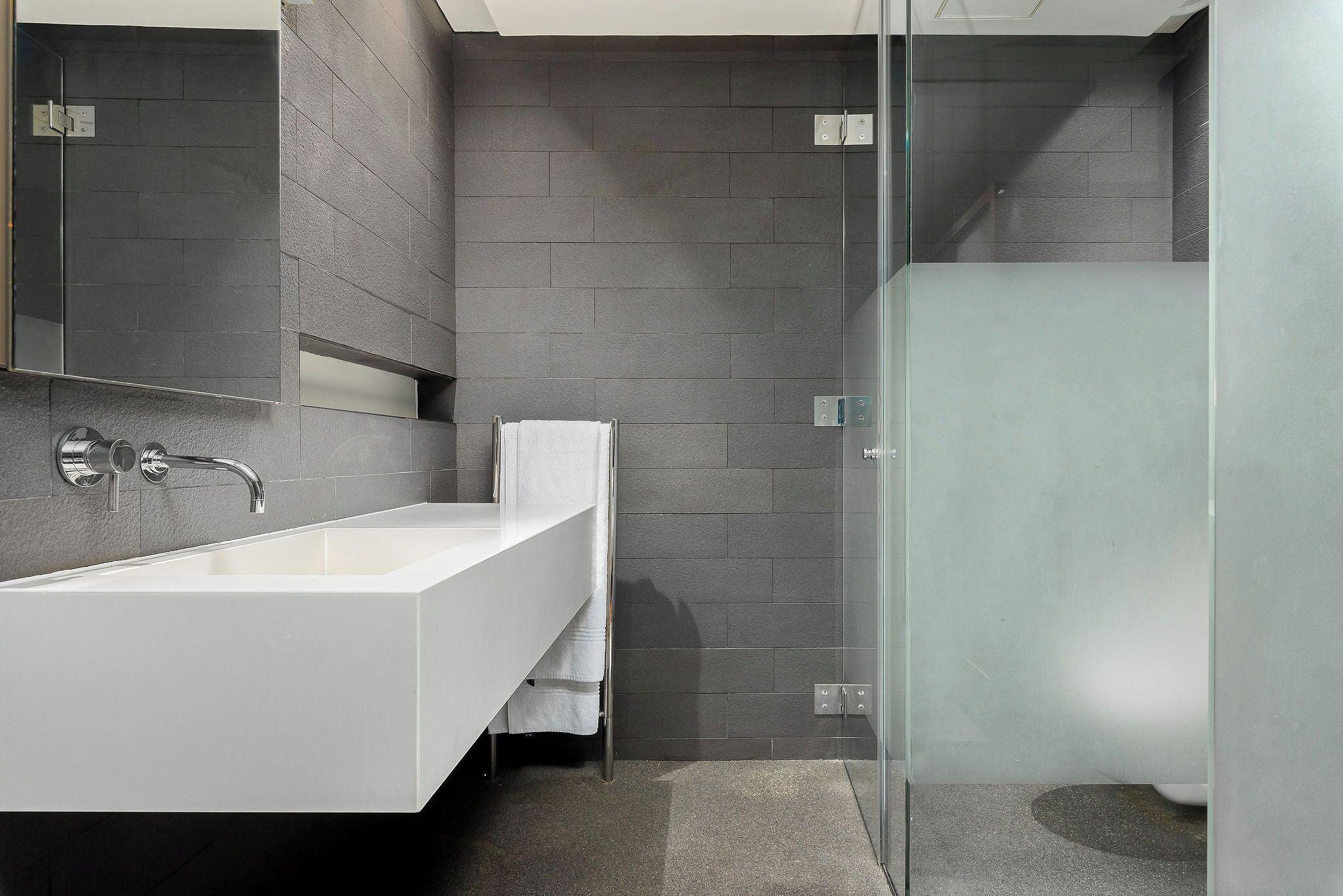 A modern bathroom with a white sink, a glass shower enclosure, and dark grey tiled walls.