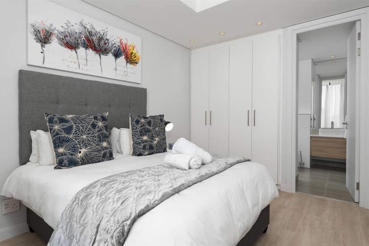 A modern bedroom with a large bed, white bedding, and decorative pillows. There is a painting above the bed and a wardrobe with sliding doors.
