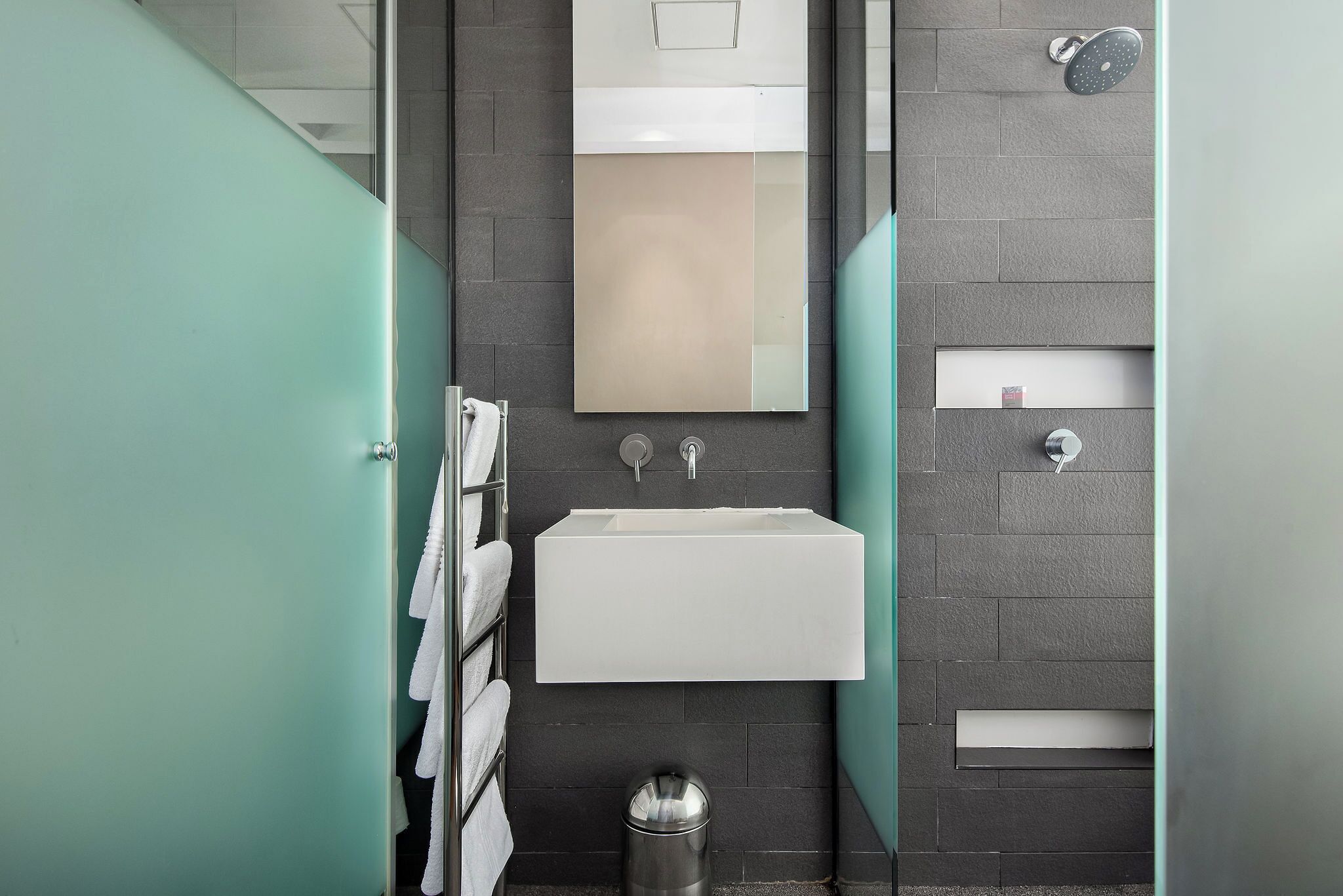 Un baño moderno con un lavabo blanco, paredes cubiertas de baldosas oscuras y una mampara de vidrio.