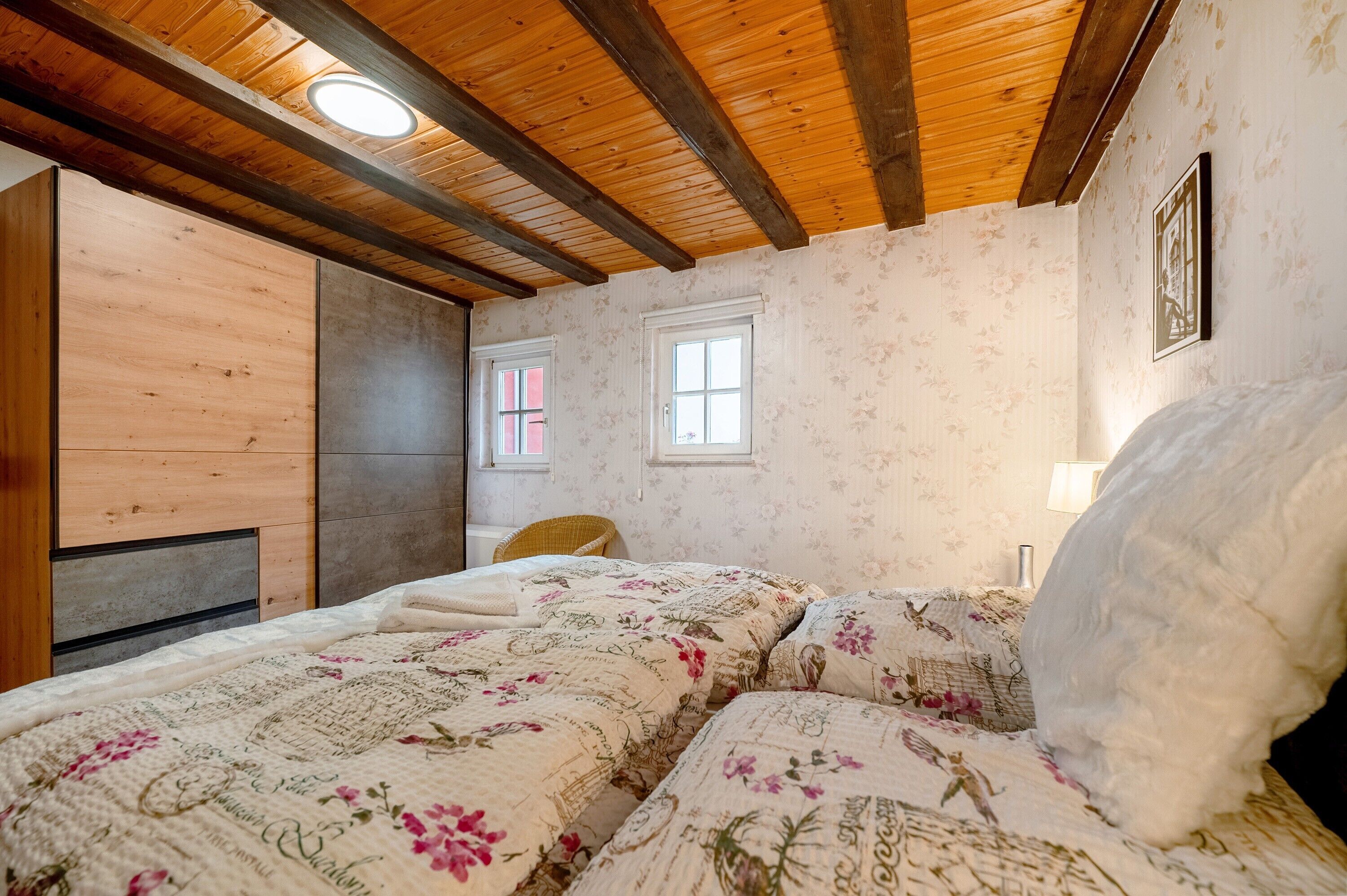 Een slaapkamer met een houten plafond, een bed met bloemenbeddengoed, een stoel en een wandlamp.