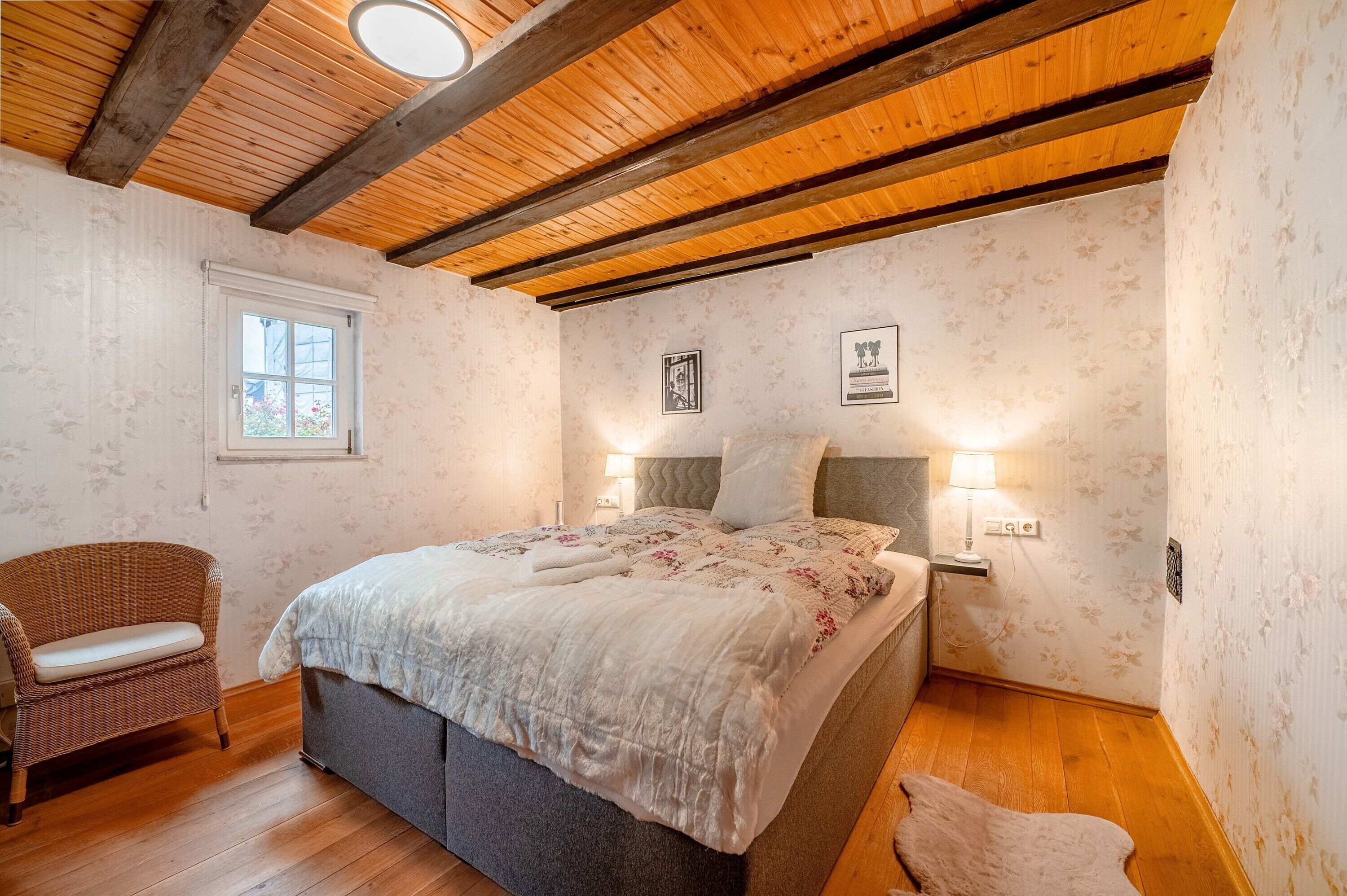 Een slaapkamer met een houten plafond, een bed met een witte dekbedovertrek, twee nachtlampjes, een rieten stoel en een raam.