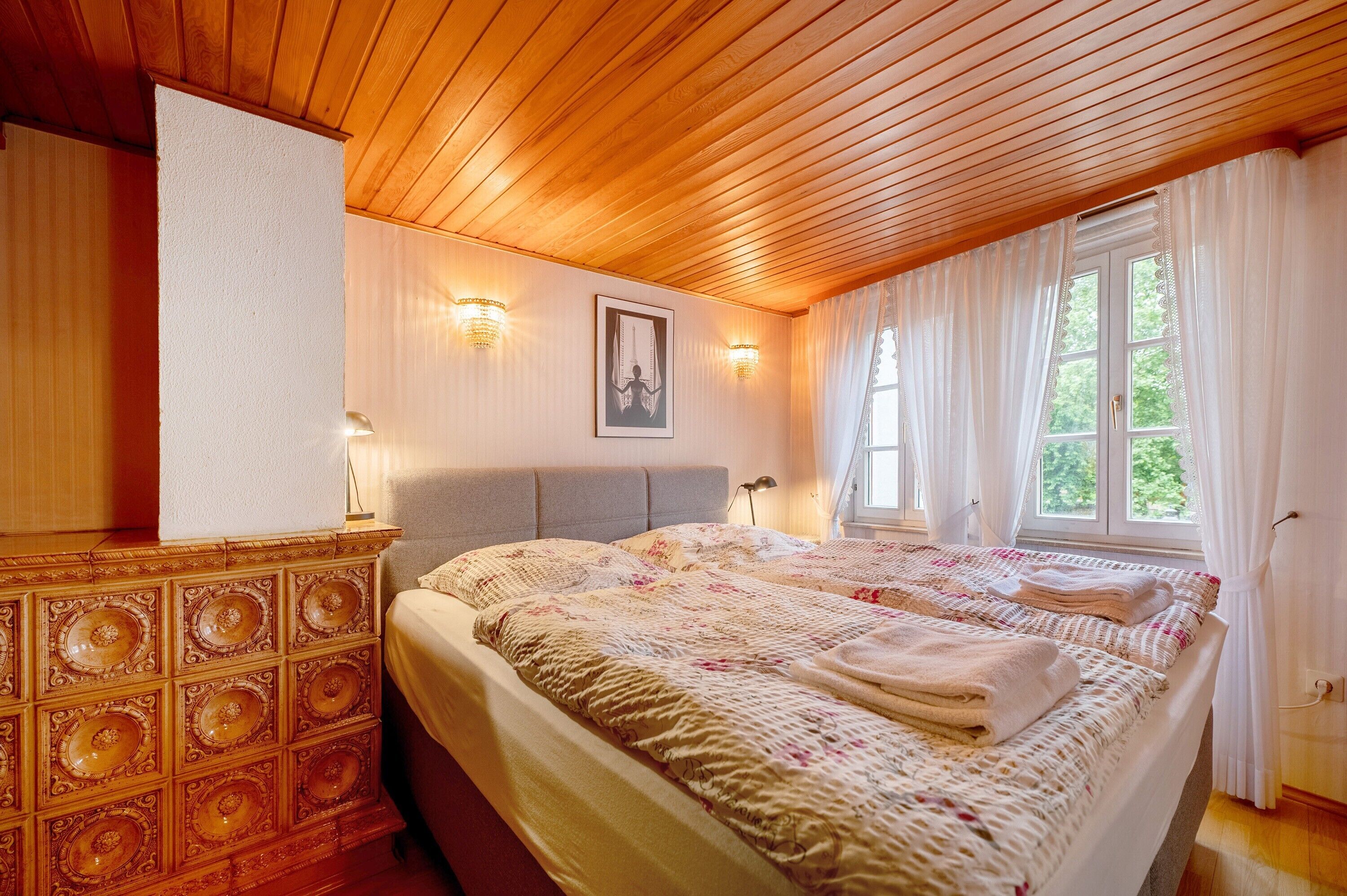 A bedroom with a wooden ceiling, a bed with a floral bedspread, a dresser, and a window with curtains.