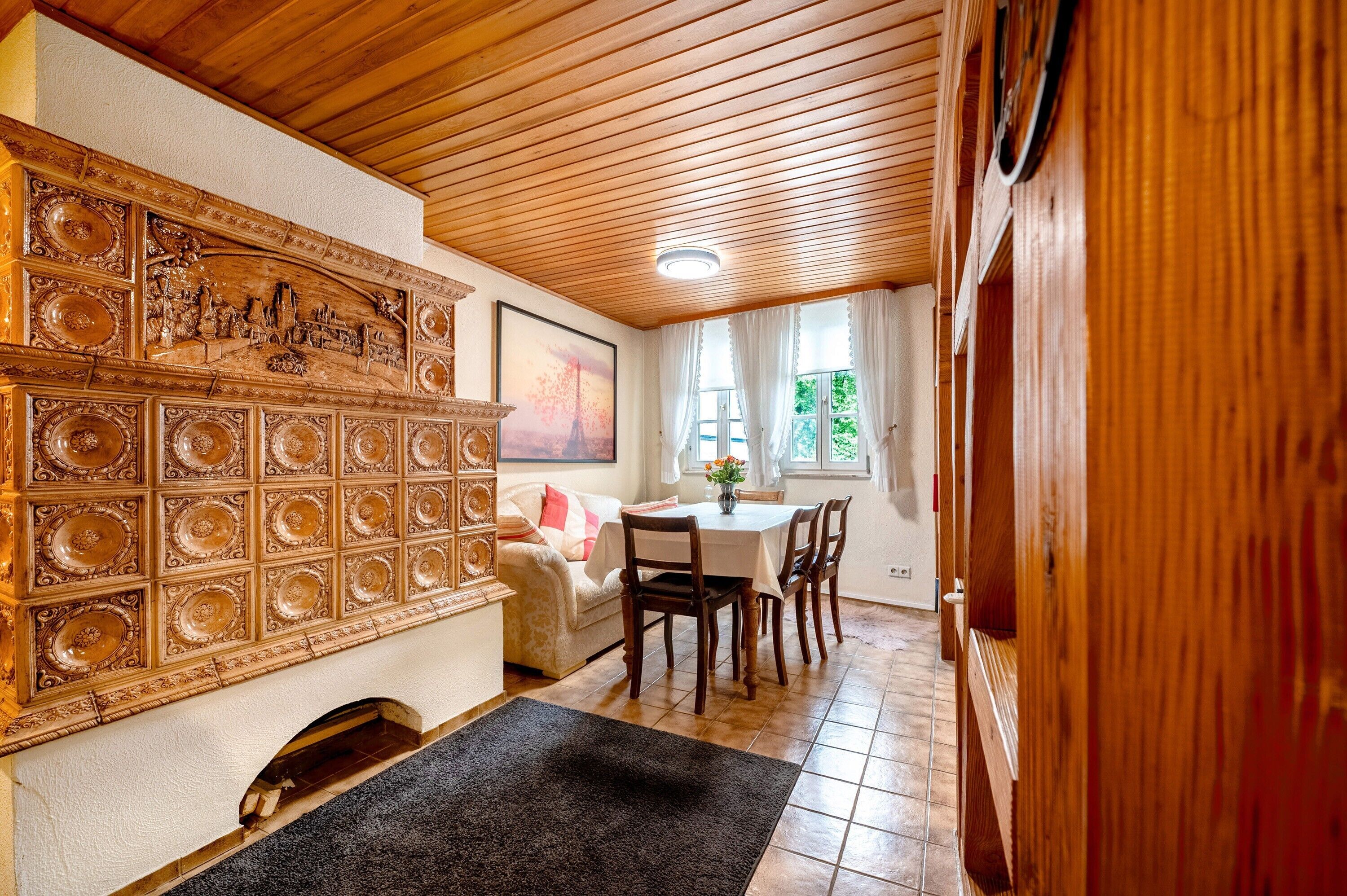 A room with a wooden ceiling, a fireplace with intricate designs, a dining area with a table and chairs, and a window with curtains.