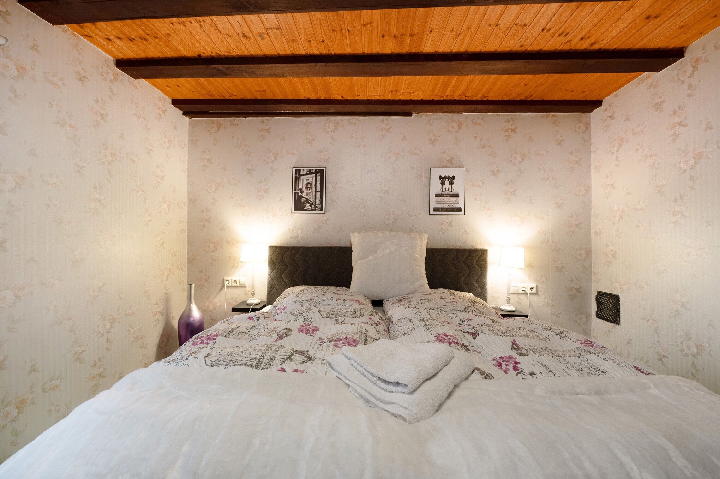 Une chambre avec un plafond en bois, un papier peint à motifs floraux, un lit avec une literie à motifs floraux, deux lampes de chevet et une table de chevet avec un vase.