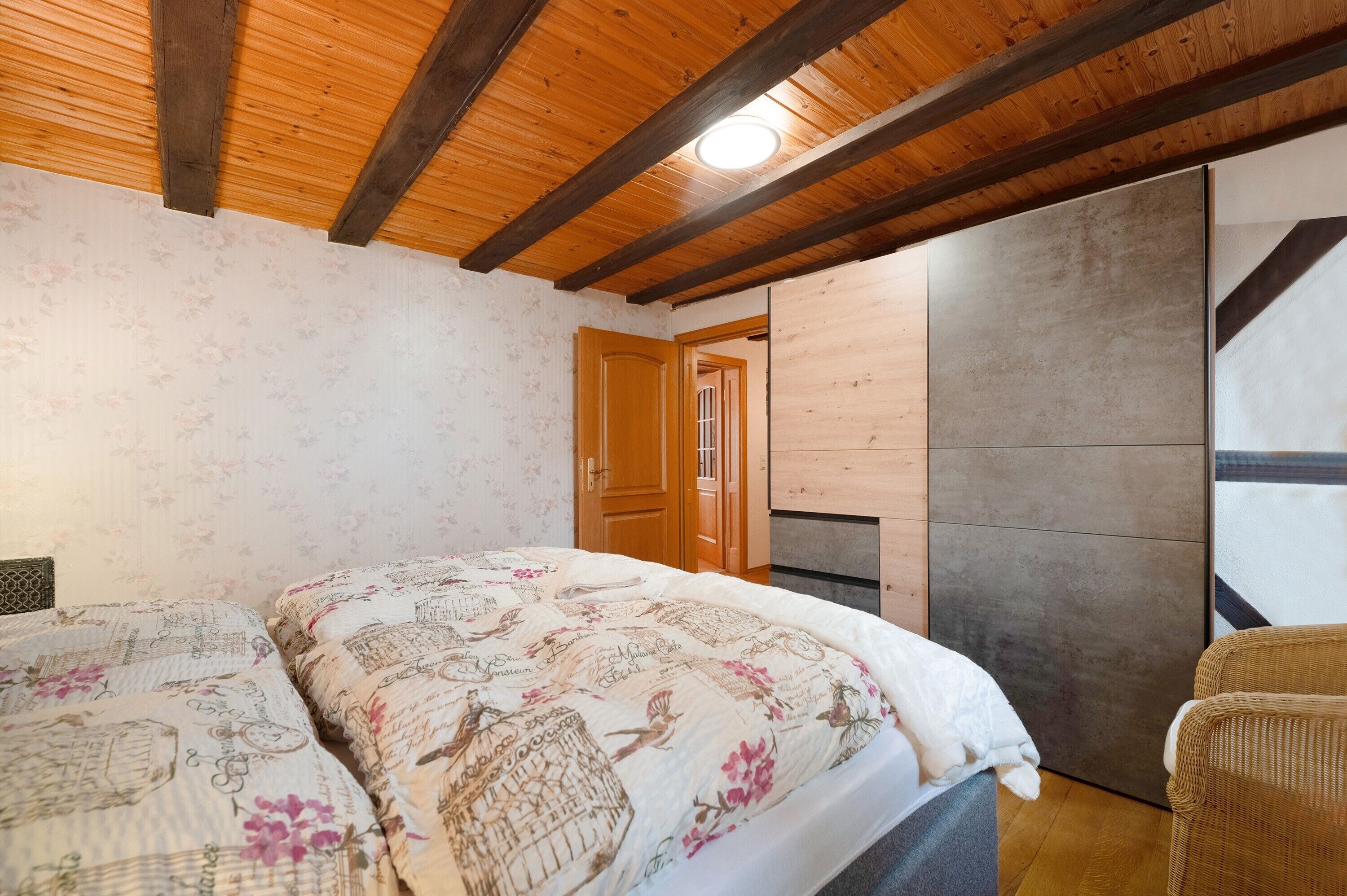 A bedroom with a wooden ceiling, two beds with floral bedding, a wooden door, and a stone wall.