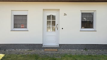 Ein weißes Gebäude mit einer weißen Tür, zwei Fenstern mit Spitzgittervorhängen und der kleinen Zahl 2 an der Wand.