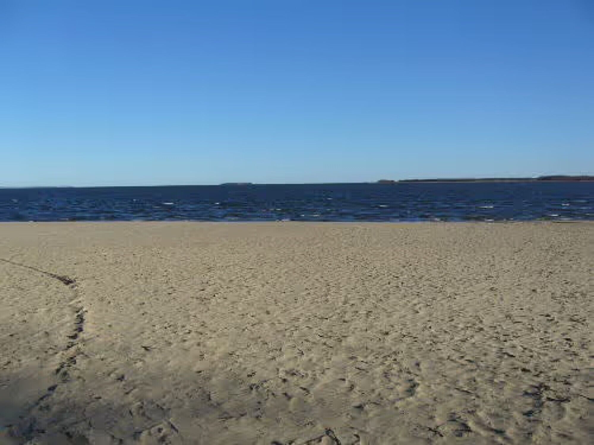 A beach with footprints leading to the water, a clear sky, and a distant landmass.