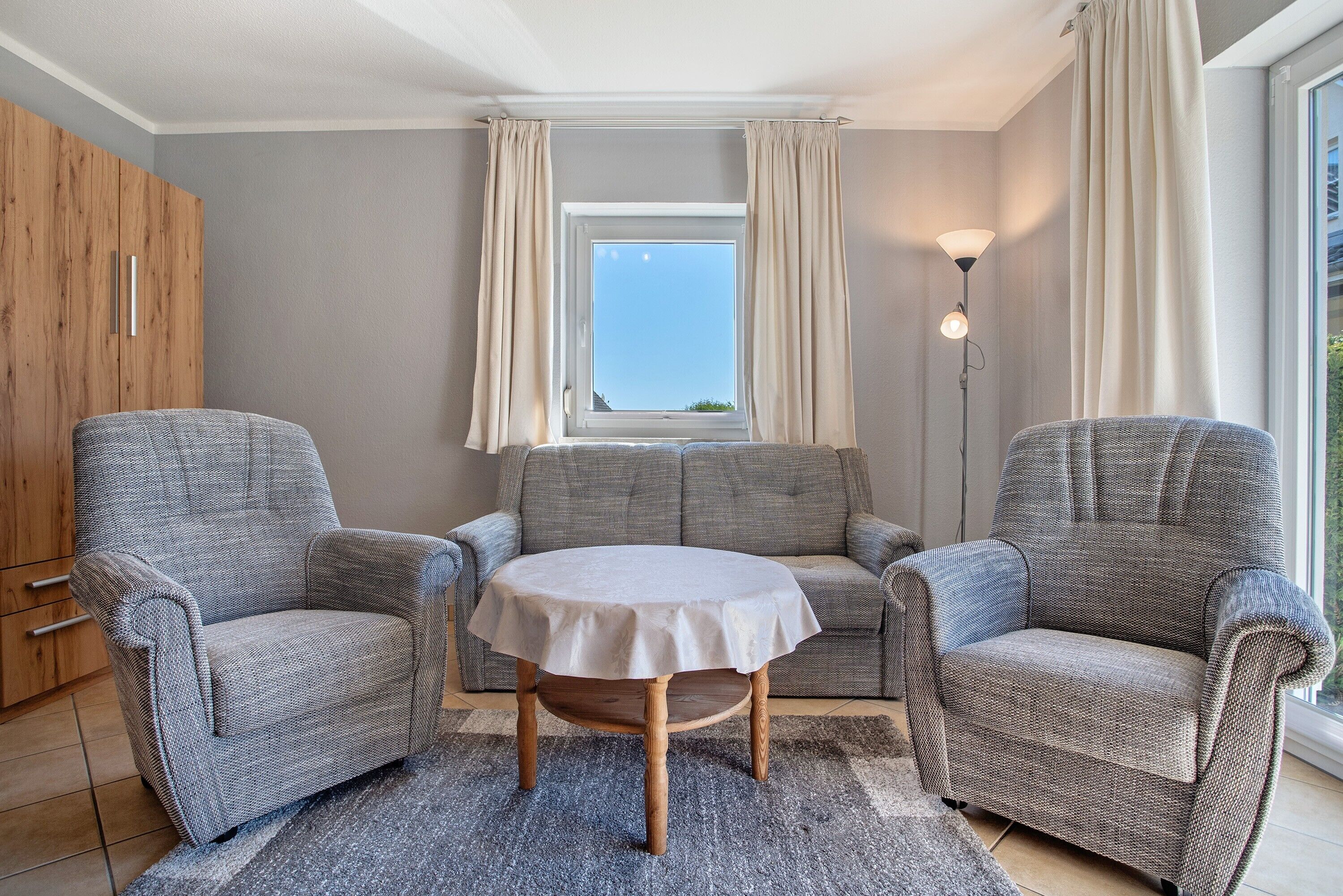 Ein Zimmer mit zwei grauen Sesseln, einem runden Tisch, einem Holzschrank und einem Fenster mit Vorhängen.