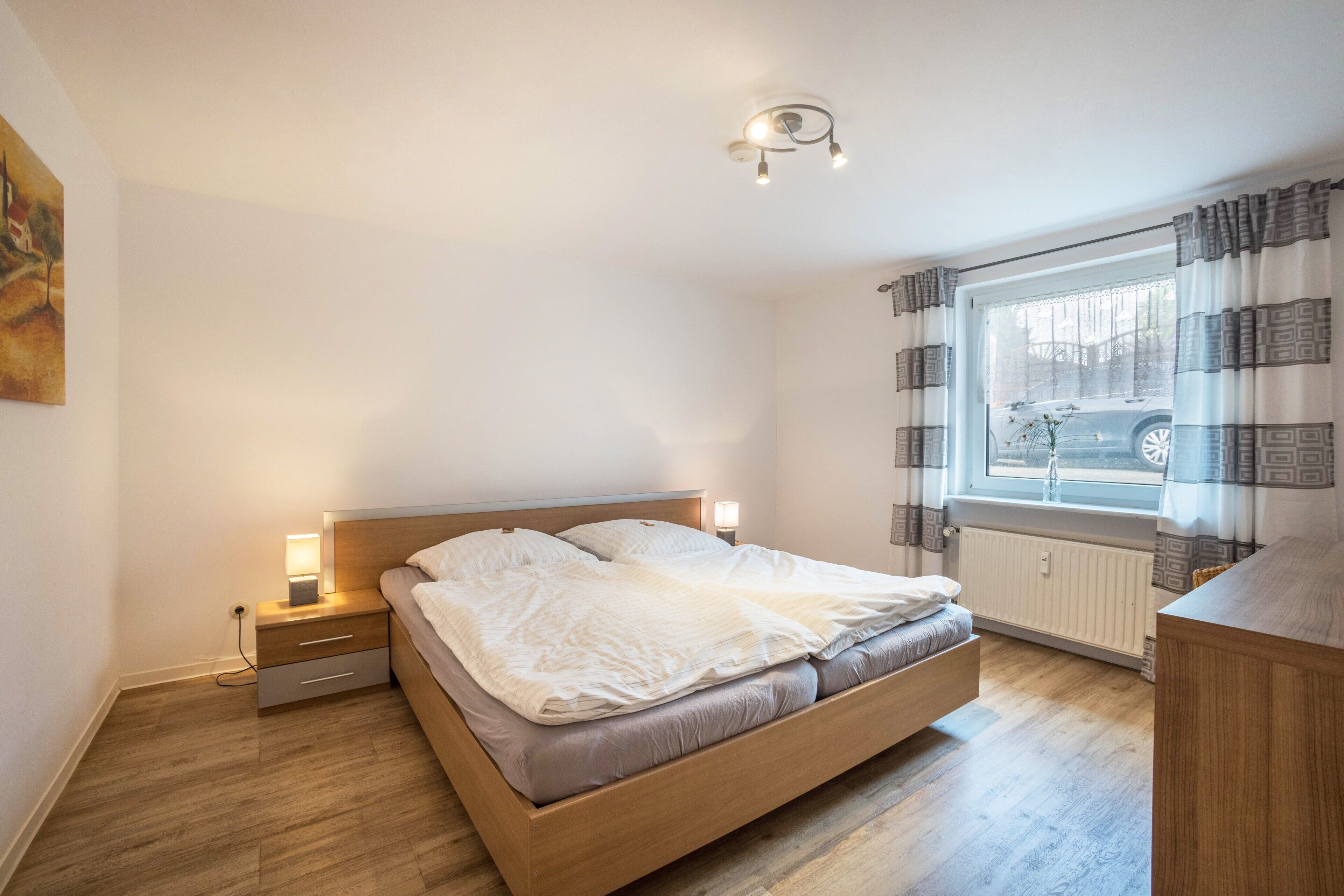 Un dormitorio con cama de madera, lámparas de noche, una ventana con cortinas y un radiador.