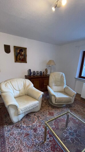 Un salón con dos sillones blancos de cuero, un tapiz estampado, un mueble de madera con adornos y una lámpara.