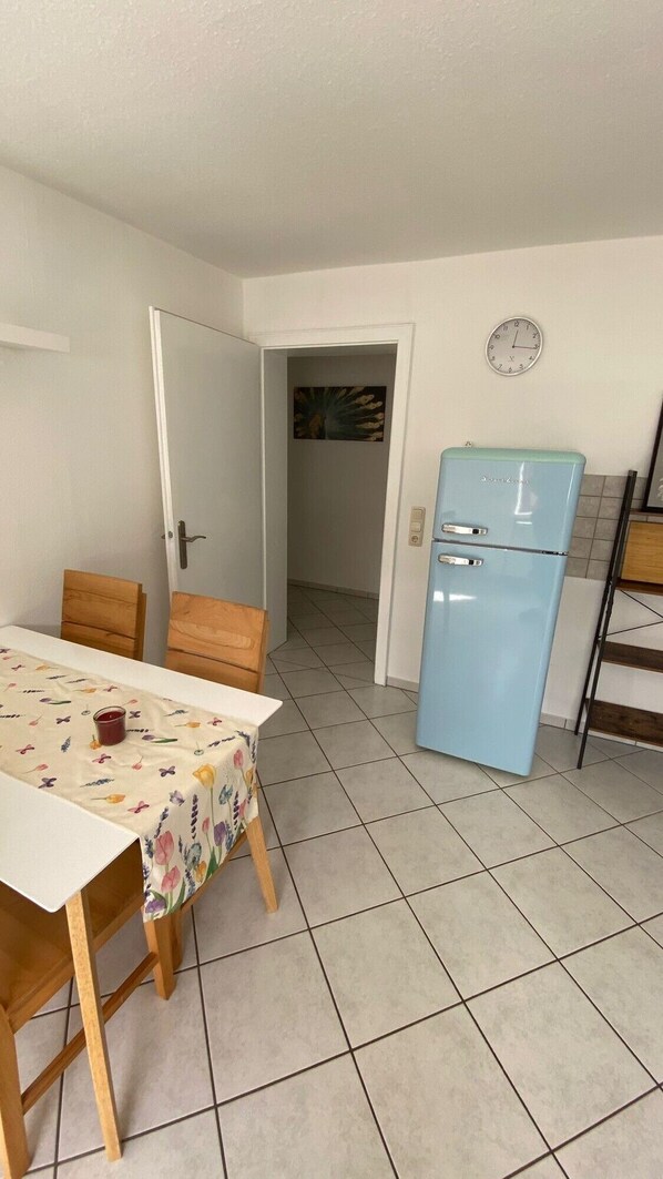 Una cocina compacta con un refrigerador azul, una mesa de comedor con mantel floreado y una puerta que da a otra habitación.