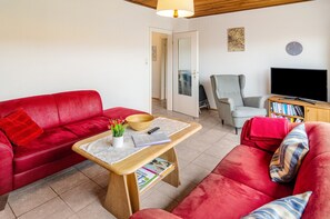 A living room with a red sofa, a wooden coffee table, a grey armchair, a bookshelf, and a television.