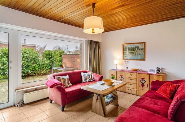 A living room with a red sofa, a wooden table, a lamp, a painting, and a view of a garden through a window.