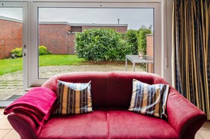 A red sofa with striped pillows, a small table, and a view of a brick wall and green hedge through a glass door.