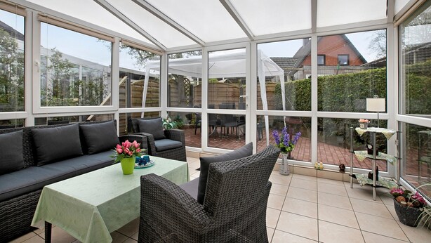 En overdækket terrasse med sofa, bord og stole, store vinduer samt udsigt til baghaven.